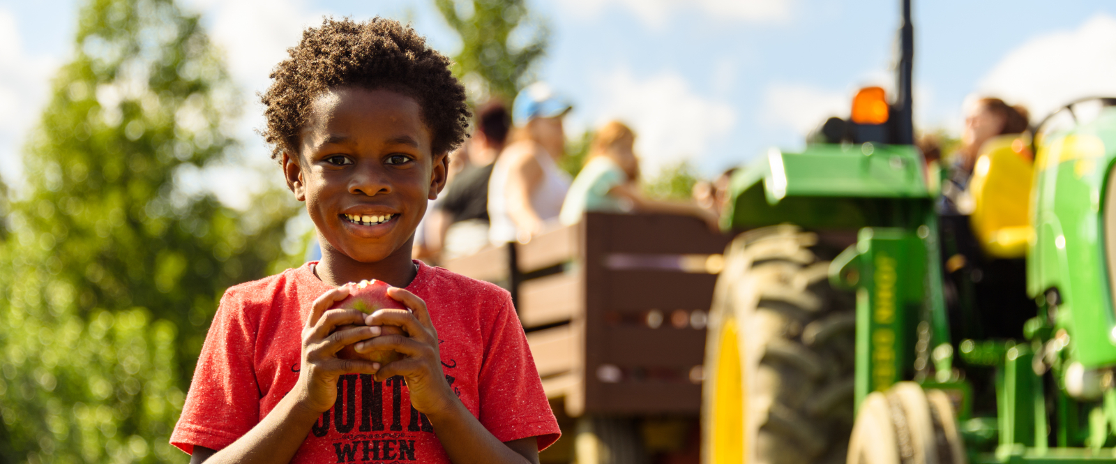 Smiling boy holding apple near tractor and wagon with passengers