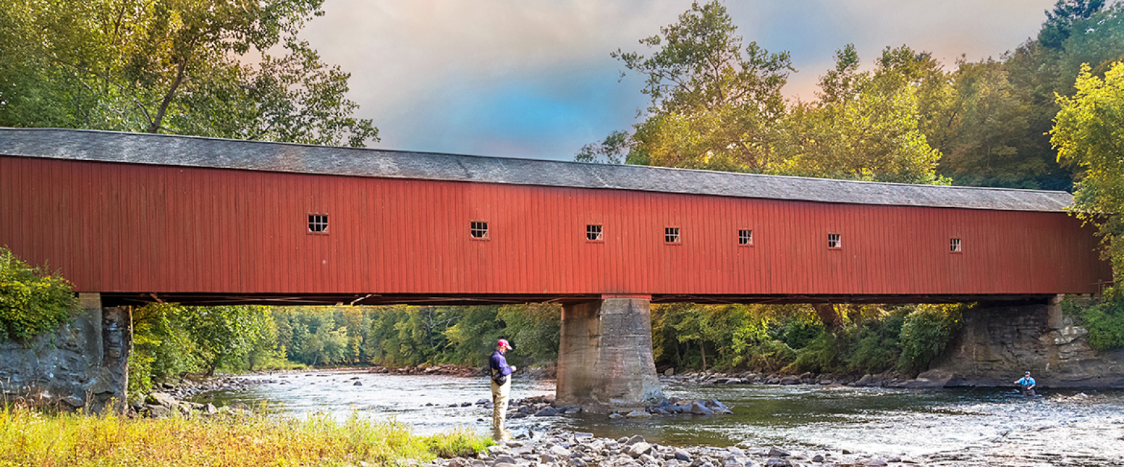 Red covered bridge over river with two people fishing nearby