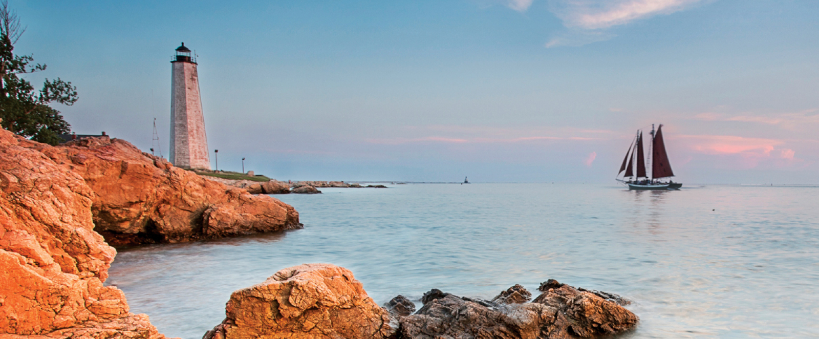 Lighthouse on rocky coast with sailboat on calm sea at sunset