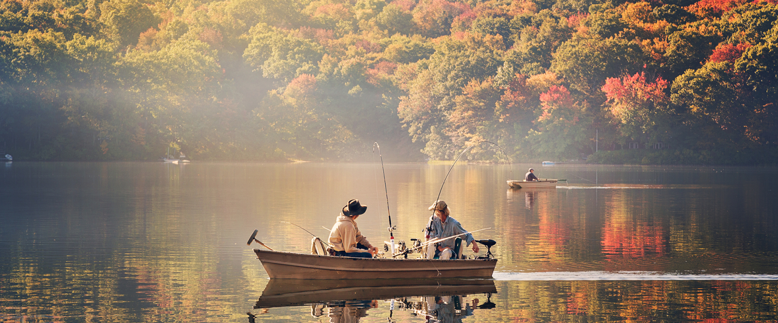 Two people fishing from a boat on a calm lake surrounded by autumn trees