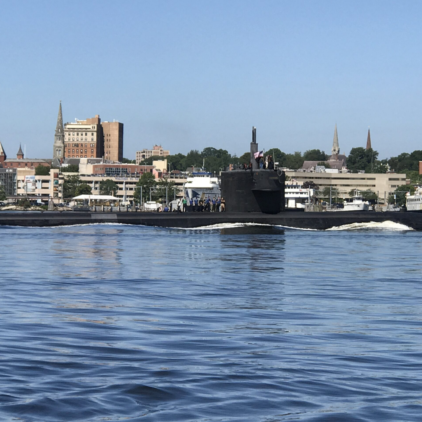 U.S. Navy submarine traveling on the Thames River with the New London waterfront and city skyline in the background.