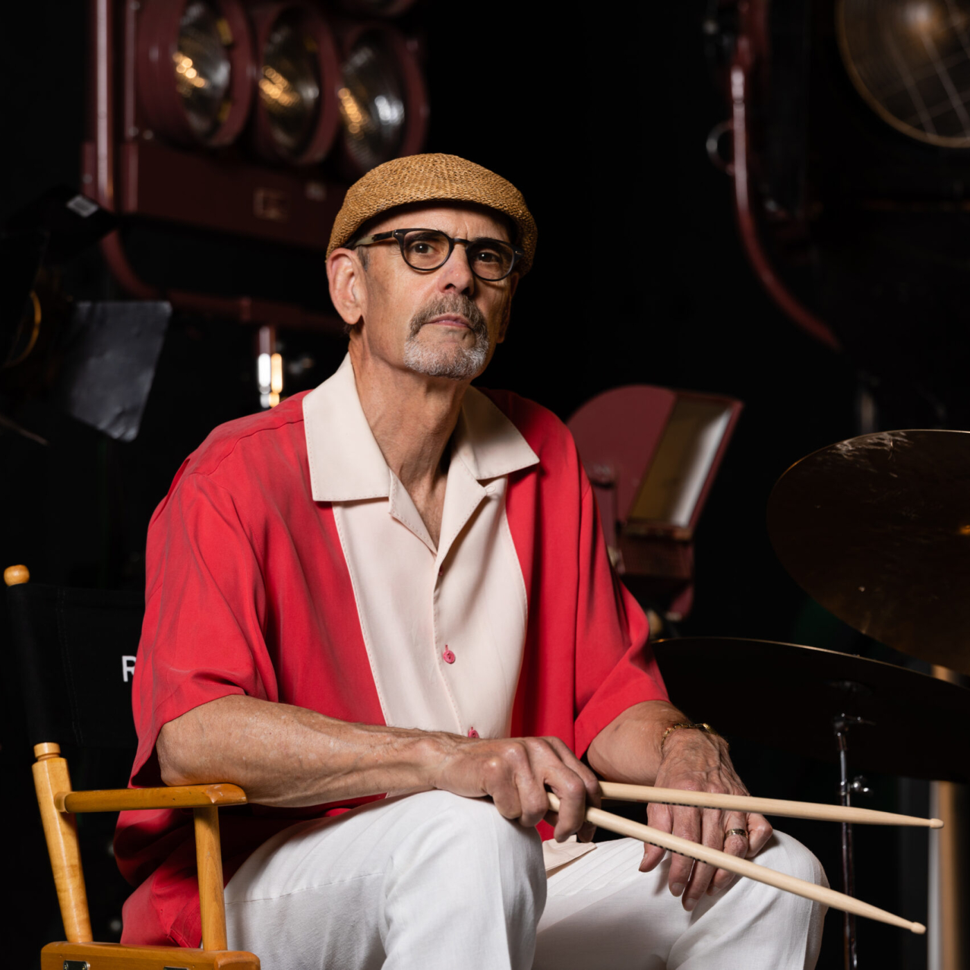 Man in red and white shirt with a tan flat cap, and white pants holding drumsticks