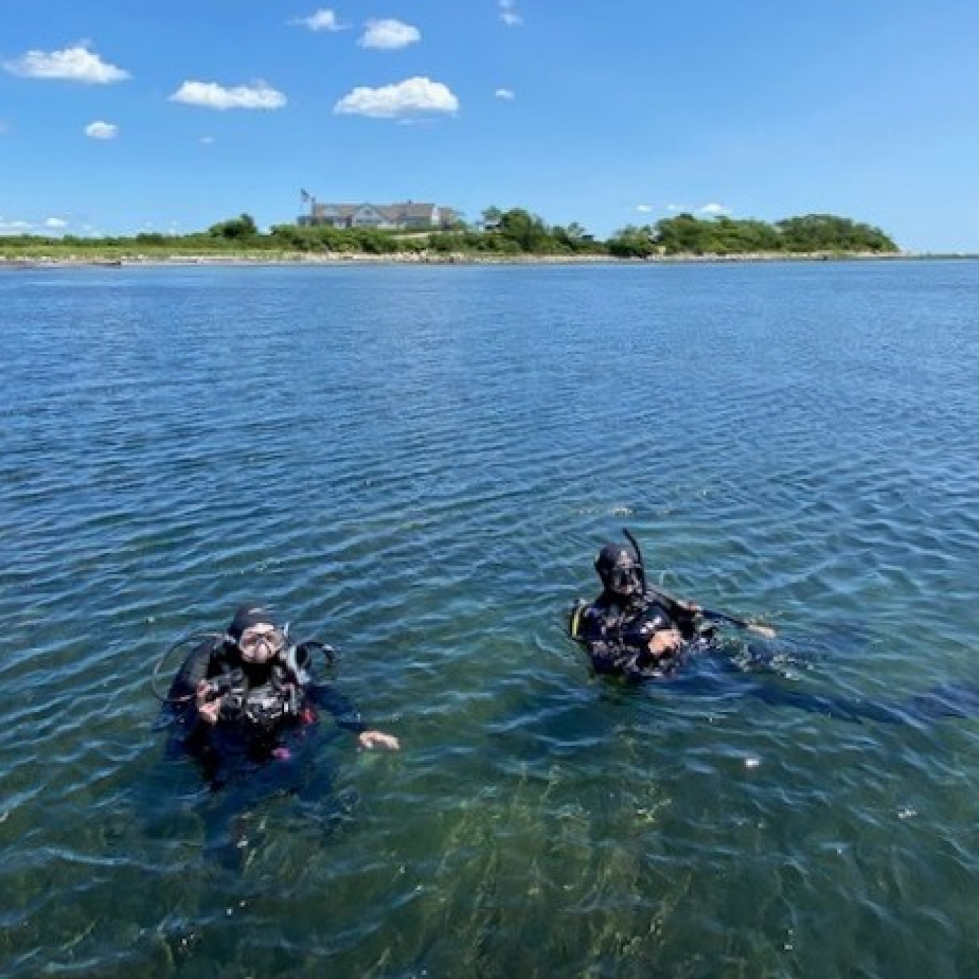 Two scuba divers floating in the water during an Eelgrass Tour at Fishers Island, with shoreline and blue sky in the background.