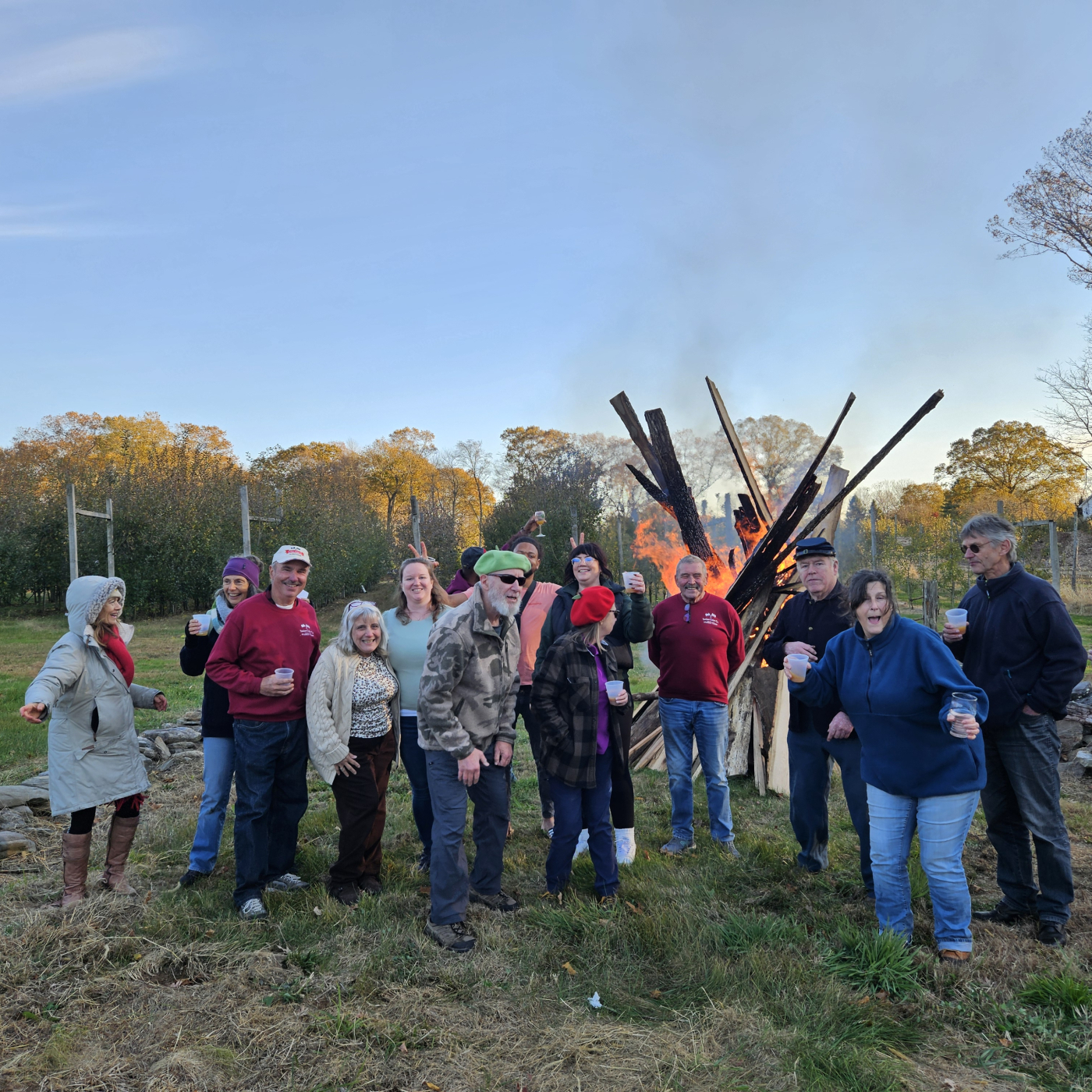 A group of people celebrating apples in an orchard, with a large fire behind them