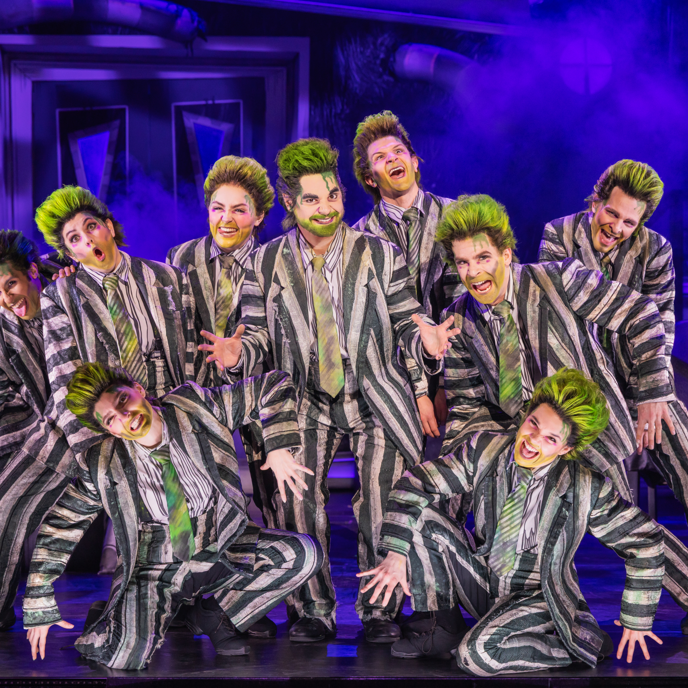 Group of male-presenting actors with green hair and striped suits on stage.