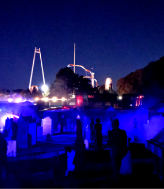 Dark haunted graveyard with glowing blue fog and amusement park rides behind