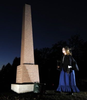 Person holding lantern near tall grave monument at night