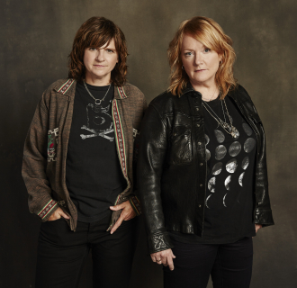 The Indigo Girls (Amy Ray and Emily Saliers) in front of a brown background