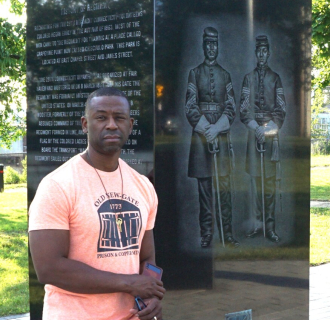 This picture is of John Mills, a Black man wearing an orange short sleeve t shirt. He is standing beside a monument that depicts two soldiers.