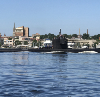 U.S. Navy submarine traveling on the Thames River with the New London waterfront and city skyline in the background.