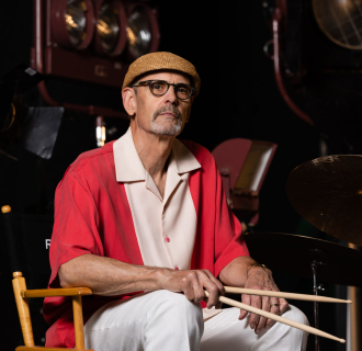 Man in red and white shirt with a tan flat cap, and white pants holding drumsticks