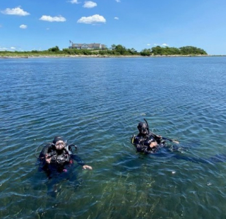 Two scuba divers floating in the water during an Eelgrass Tour at Fishers Island, with shoreline and blue sky in the background.