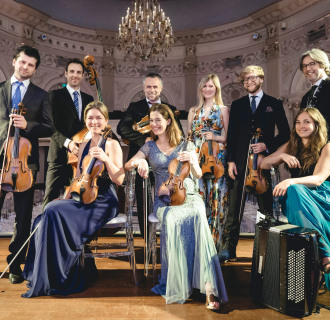 Group portrait of a chamber orchestra standing and seated in an ornate concert hall