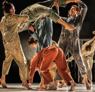 A group of dancers in coveralls perform dynamic movements