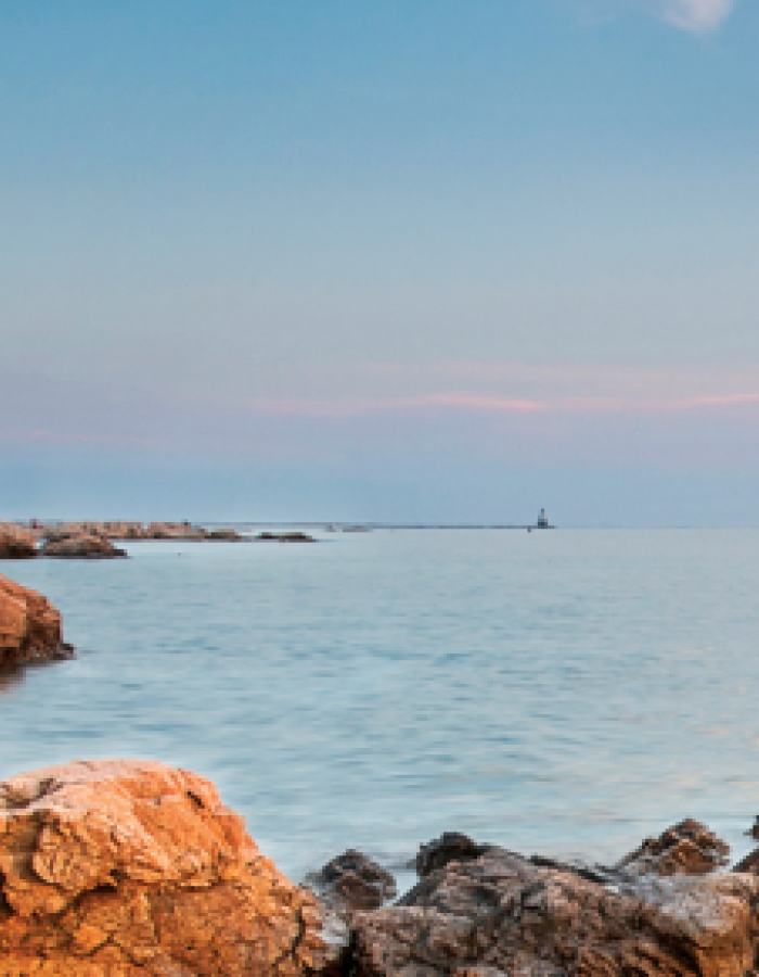 Lighthouse on rocky coast with sailboat on calm sea at sunset