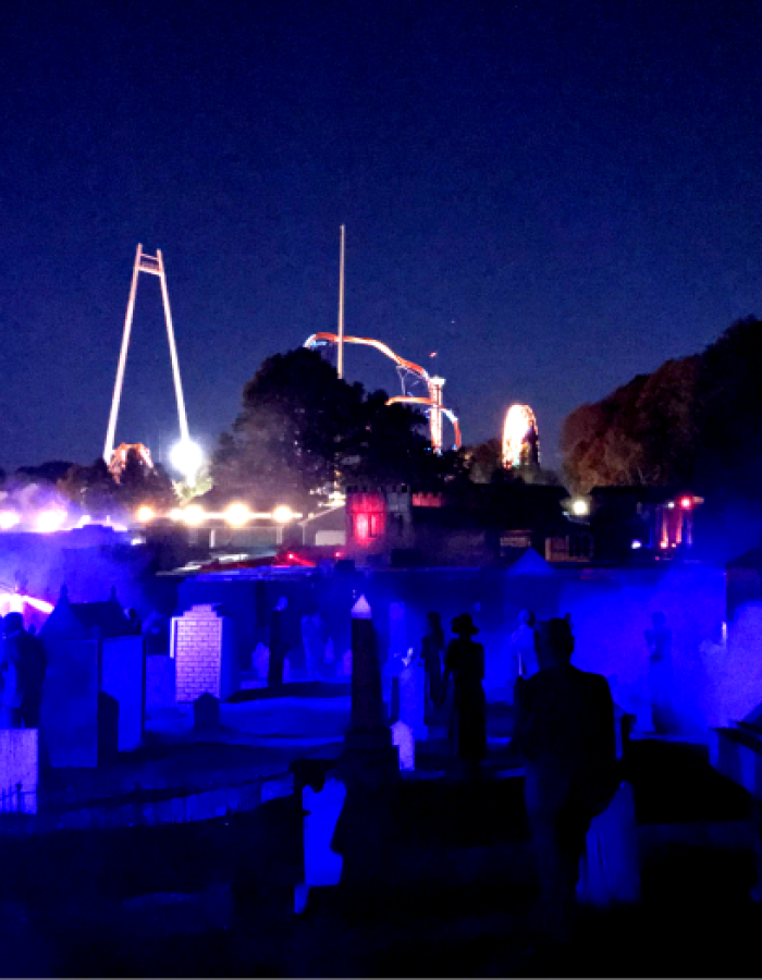 Dark haunted graveyard with glowing blue fog and amusement park rides behind