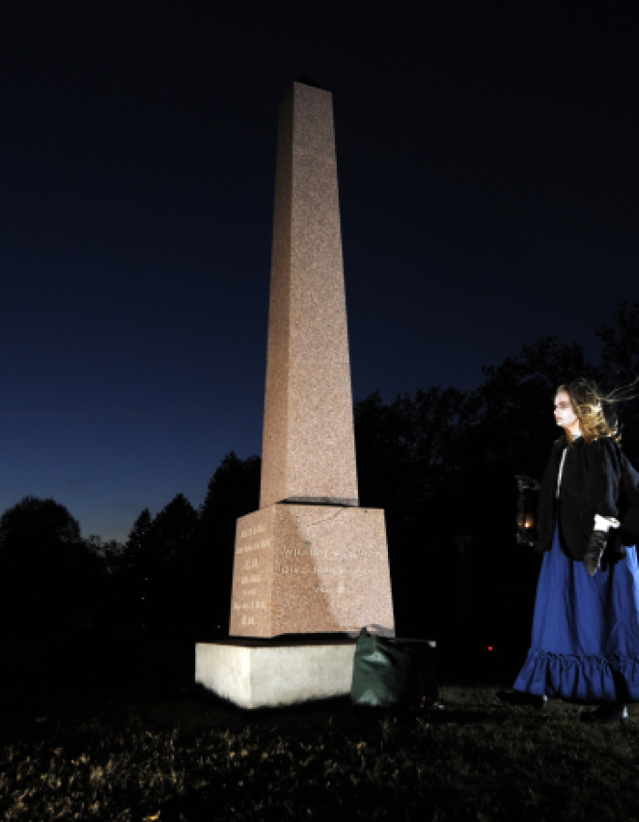 Person holding lantern near tall grave monument at night