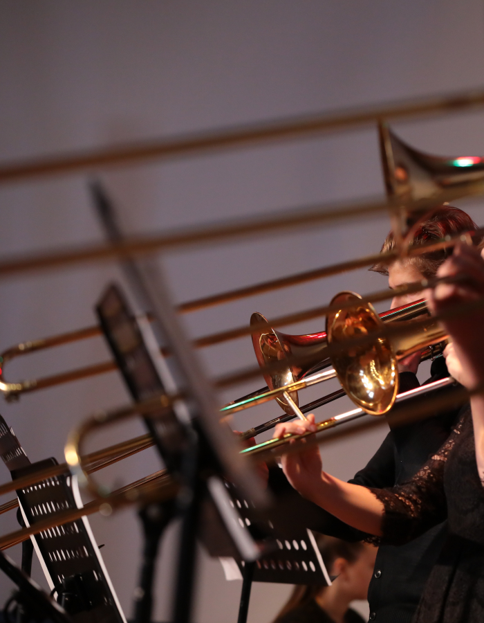 Brass musicians performing with trombones and trumpets on stage, viewed from the side.