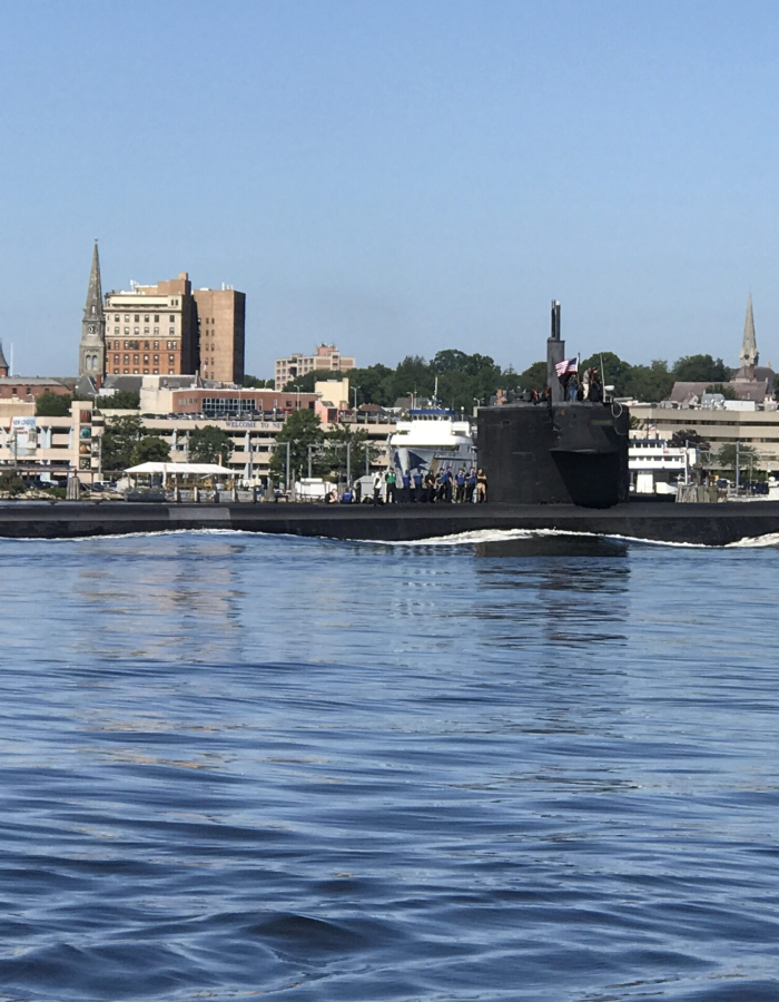 U.S. Navy submarine traveling on the Thames River with the New London waterfront and city skyline in the background.