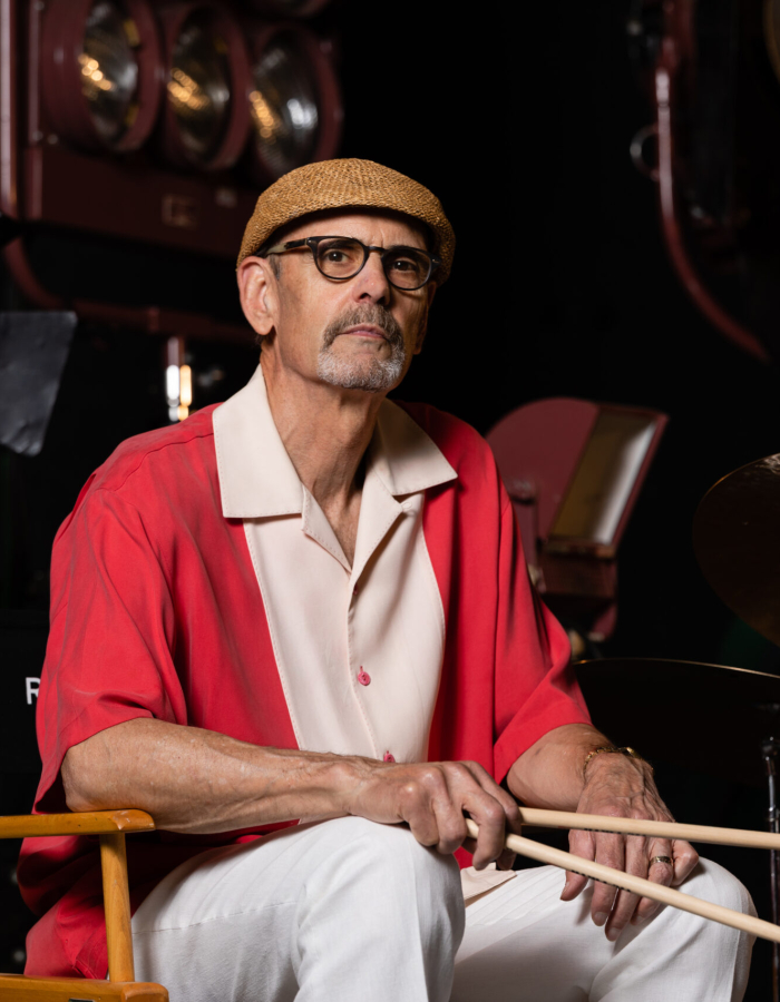 Man in red and white shirt with a tan flat cap, and white pants holding drumsticks