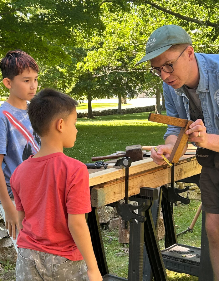 Two children watch as Andrew Rowand from the Eric Sloane Museum explains how to use a tool called a square