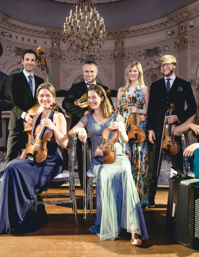 Group portrait of a chamber orchestra standing and seated in an ornate concert hall