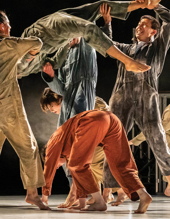 A group of dancers in coveralls perform dynamic movements