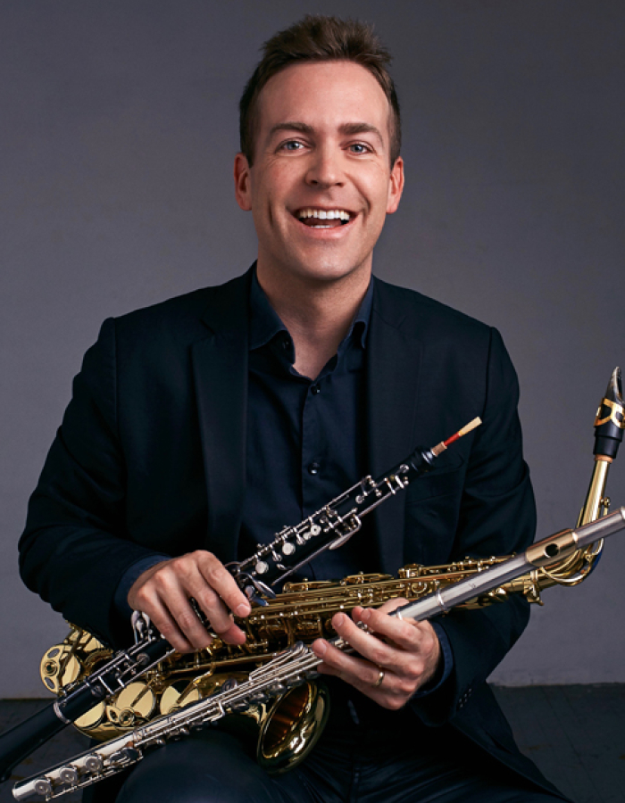 Man in a suit smiling and holding a saxophone, flute, and oboe