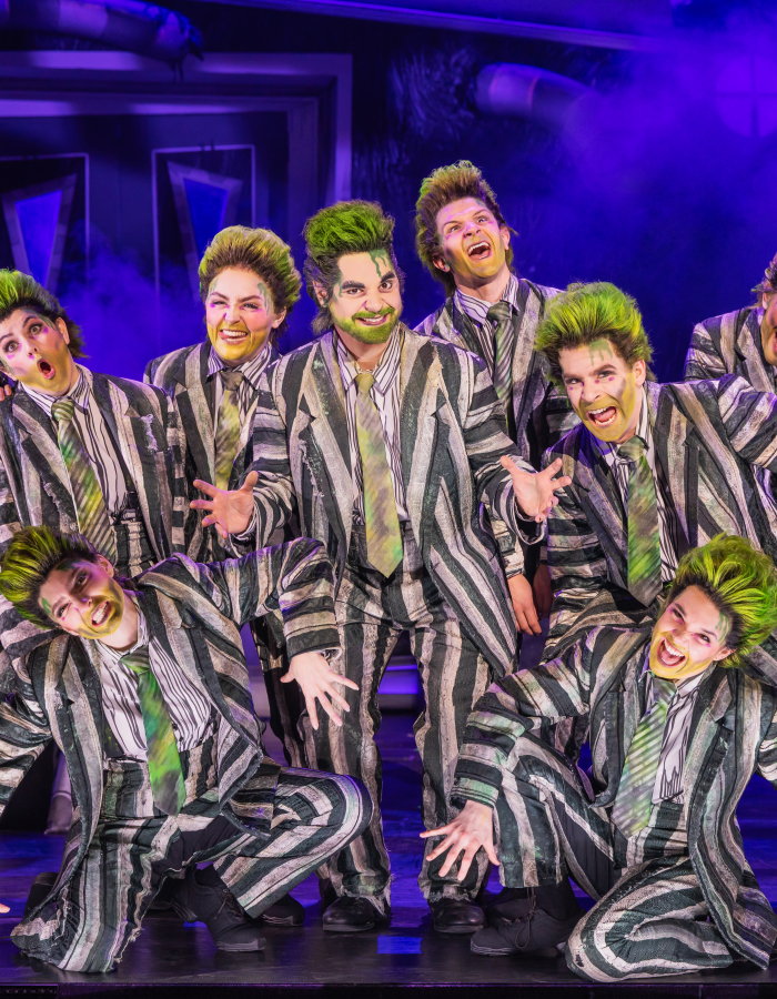 Group of male-presenting actors with green hair and striped suits on stage.