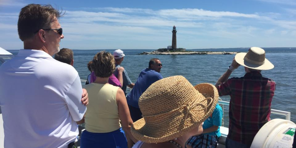 Tourists on boat viewing lighthouse on rocky island under blue sky