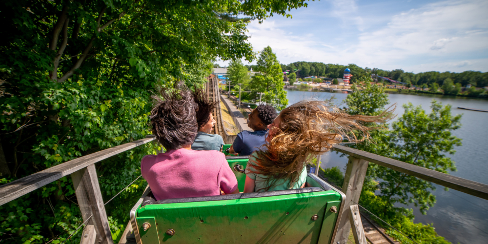 Four people with windblown hair riding roller coaster near water and trees