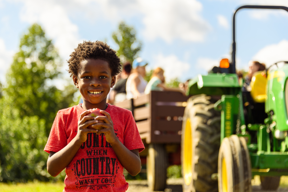Smiling boy holding an apple with a tractor and wagon in background