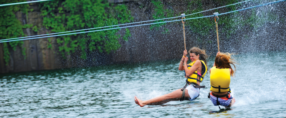 Two people in life jackets riding zipline over water splashing