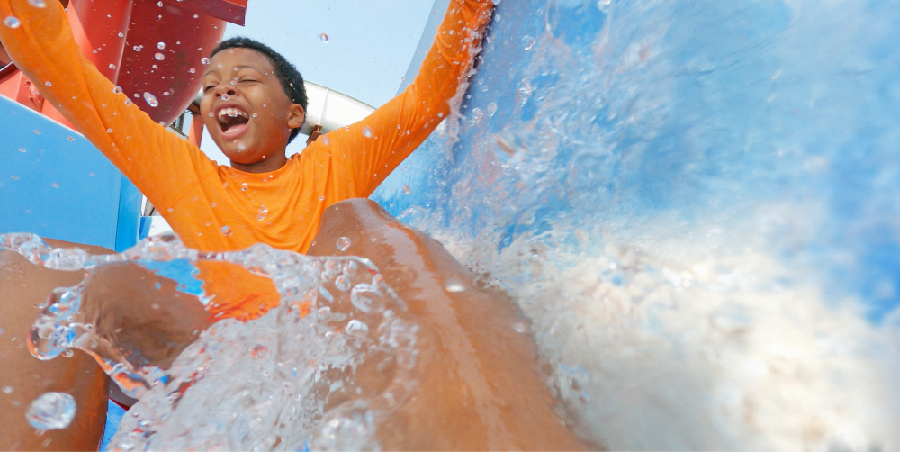 Boy enjoying water slide wearing orange shirt with arms raised