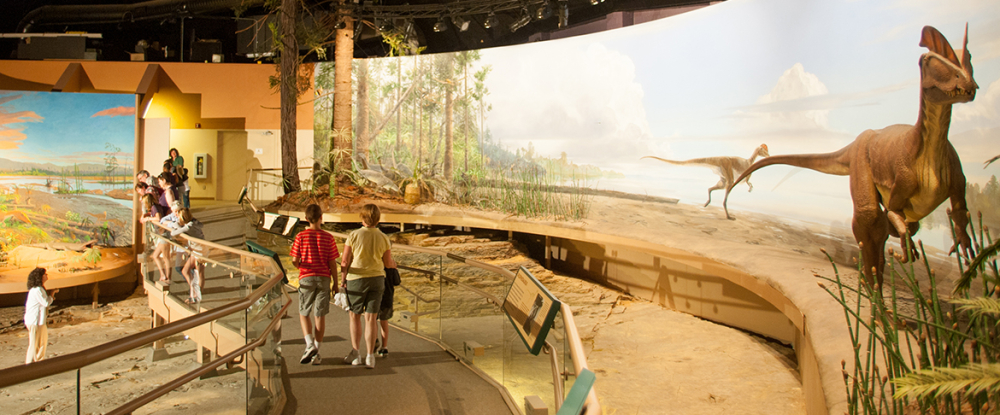 Visitors walk through dinosaur exhibit with life-sized dinosaur models and forest backdrop