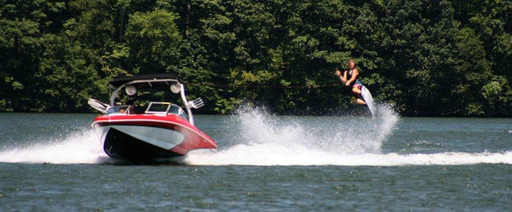 Person wakeboarding behind a red and white boat on a lake with trees