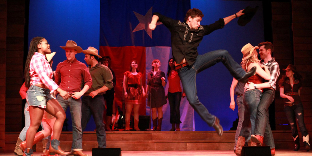 Actors performing a lively country dance onstage with Texas flag backdrop