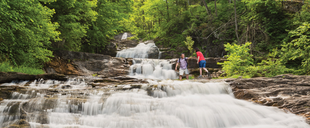 Two hikers helping each other beside cascading waterfall in forest