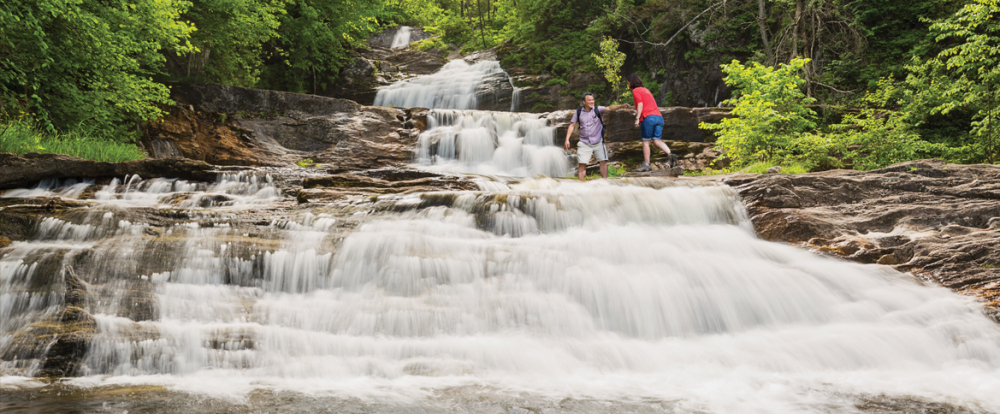 Two hikers on rocks beside cascading multi-tiered Kent Falls waterfall