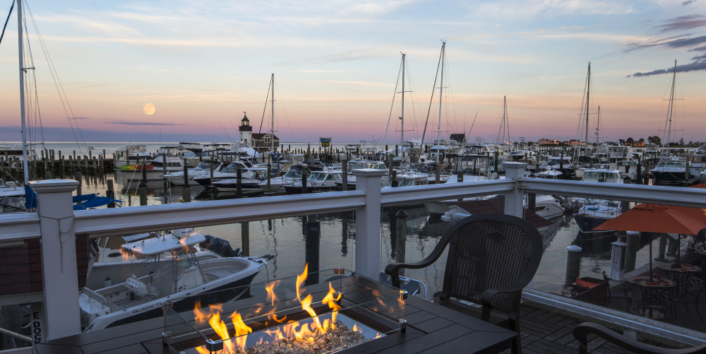 Marina with boats and lighthouse at sunset, fire pit and chairs in foreground