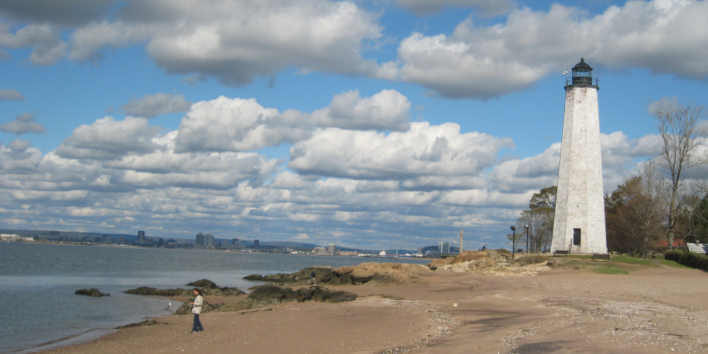 White lighthouse on rocky beach with cloudy sky and city skyline in distance