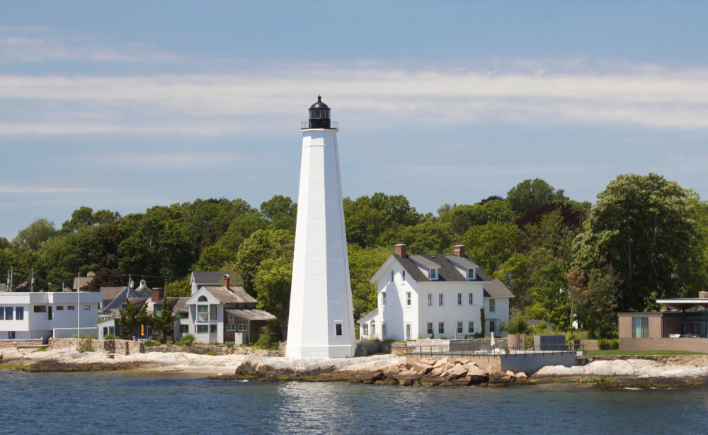 White New London Harbor Lighthouse on rocky shoreline with houses and trees