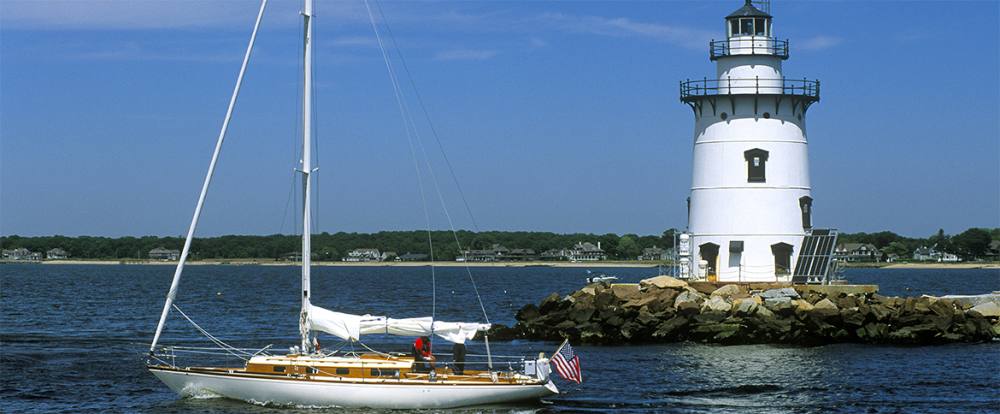 Sailboat near a white lighthouse on rocky shore with houses in background