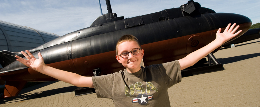 Boy with glasses in front of a black and orange submarine outdoors