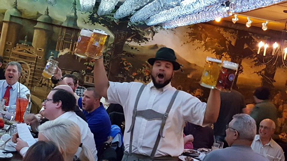 Man in traditional outfit holding four large beer mugs in busy beer hall