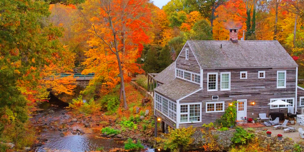 Gray wooden house by river with colorful autumn trees and stone bridge