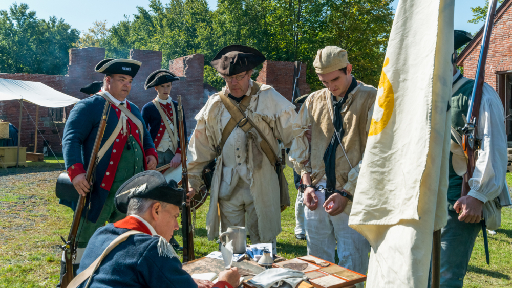 Costumed interpreters in the prison yard. One is dressed as an officers and is sitting while he documents the prisoner being presented to him in handcuffs. 