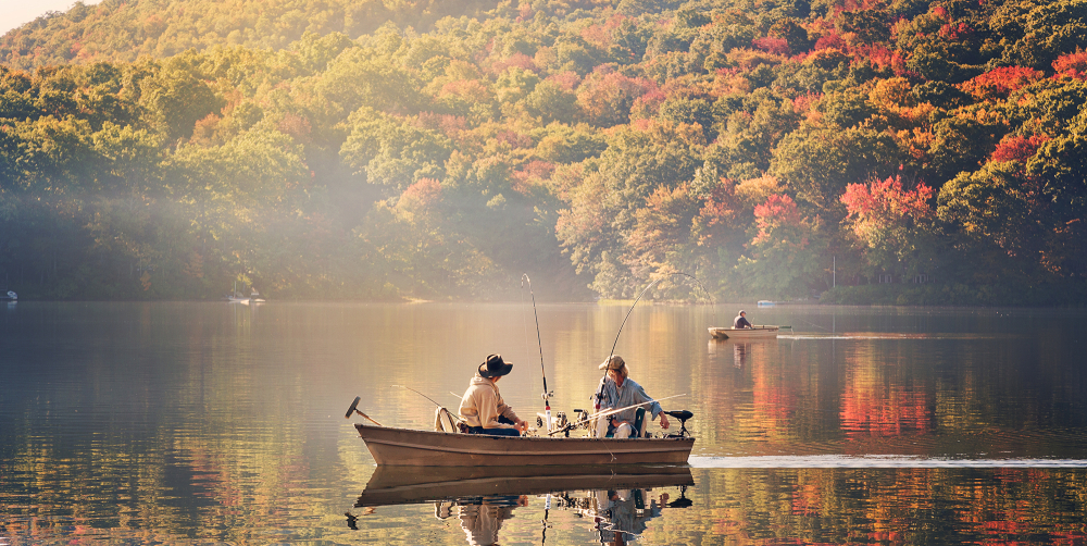 Two people fishing from a boat on a calm lake with autumn trees
