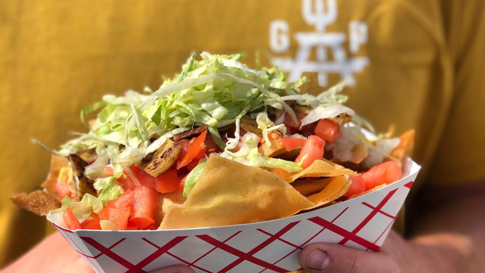 Person holding nachos topped with lettuce, tomatoes, and grilled meat