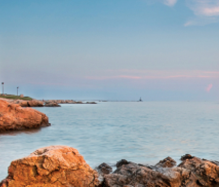 Lighthouse on rocky coast with sailboat on calm sea at sunset